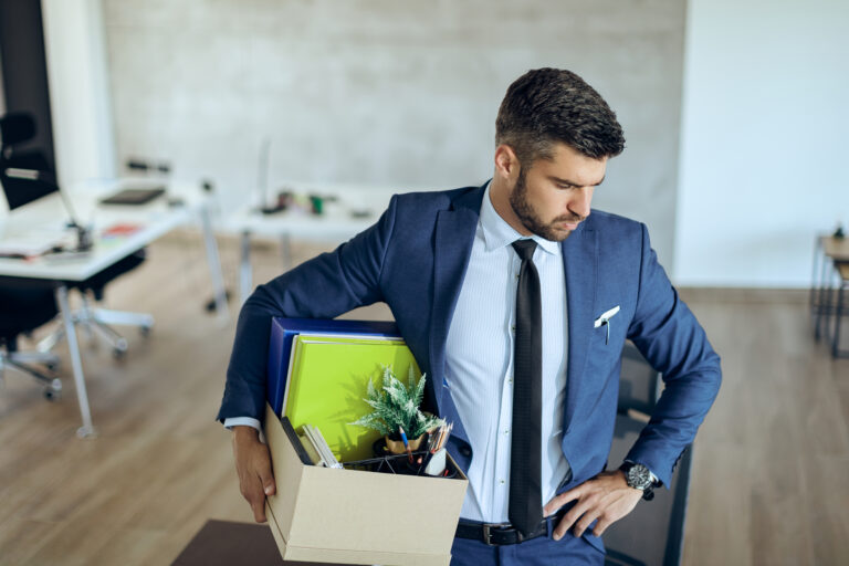 Ein Mann im Anzug trägt eine Kiste in einem Büro und erlebt dabei Chef-Stress.